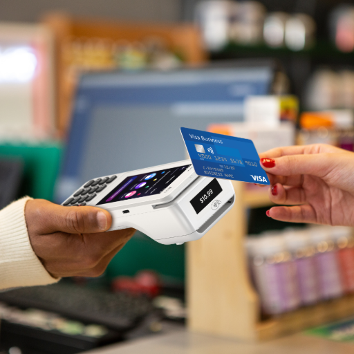 L'image montre une scène de paiement sans contact à un comptoir de magasin. La main d'un client, avec du vernis à ongles rouge, tient une carte bancaire Visa bleue au-dessus d'un terminal de paiement blanc moderne, tenu par la main du commerçant. En arrière-plan, un écran de caisse est visible. Le texte à droite, titré 'Comment choisir son terminal de paiement ?', explique qu'il est essentiel de comprendre les différents types de TPE pour savoir lequel correspond à quelle activité. Il est conseillé de se renseigner à l'avance pour éviter d'investir dans un terminal 'qui n'est pas adapté à votre commerce' en évaluant ses besoins et son budget. Le texte indique que E-MONÉTIQUE aide à faire ce choix en expliquant les 3 catégories de TPE existantes : 'Les terminaux de paiements fixes', 'Les terminaux de paiements mobiles' et 'Les terminaux de paiements portables'. comment choisir son terminal de paiement ?