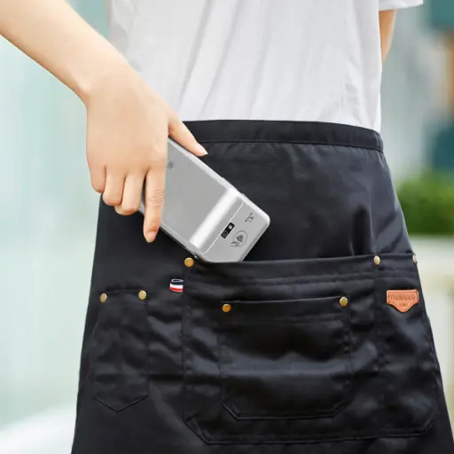 L'image montre une personne, vue de dos, portant un tablier de service noir. Sa main glisse le terminal de paiement mobile PAX A77 dans la poche arrière de son tablier, illustrant sa portabilité. Le texte à droite, titré 'PAX A77 : Pour quel secteur d'activité ?', le décrit comme l'outil parfait pour les professionnels ayant besoin d'encaisser en dehors d'un point de vente fixe, grâce à sa grande autonomie et sa robustesse pour une utilisation intensive. Le texte liste ensuite les secteurs idéaux : 1. L'hôtellerie et la restauration, pour un encaissement à table rapide. 2. Le commerce de détail, pour réduire les files d'attente en permettant d'encaisser directement dans les rayons. 3. Les artisans et les services à domicile, pour accepter des paiements sécurisés directement chez le client. 4. La vente itinérante, comme sur les marchés ou lors d'événements, où sa batterie haute capacité assure une fiabilité de paiement tout au long de la journée.